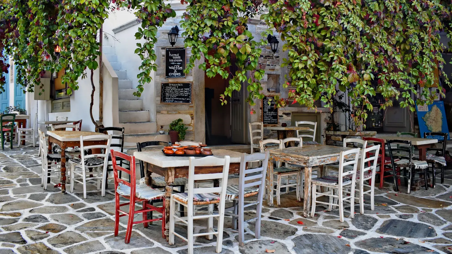 A village taverna on Naxos with mismatched wooden tables and red and white chairs on a stone-paved square, shaded by a canopy of vine leaves, a clay pot tray on one table and a map of the island on the wall